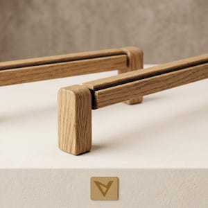 May include: Two wooden cabinet pulls with a natural wood grain finish. The pulls have a rectangular design with a black accent and are mounted on a white surface. A small, gold-colored tag with a triangle logo is in the foreground.
