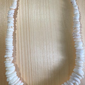 May include: A necklace composed of numerous small, irregular white shell discs strung together. The beads gradually increase in size towards the centre of the strand. The necklace is secured with a silver-toned metal lobster claw clasp and small metallic accent beads at the ends.