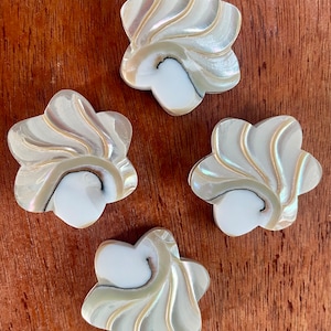 May include: Four iridescent, flower-shaped shell embellishments. Each has a white centre and swirling, pearlescent petals with hints of gold and black. The objects are arranged on a brown wooden surface.