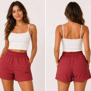 May include: A white spaghetti strap crop top paired with high-waisted, red shorts. The shorts have an elastic waistband and pockets. The outfit is casual and suitable for warm weather. The model is wearing a gold necklace.