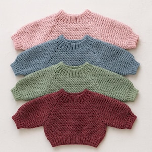 May include: Four baby sweaters in a row, each with a textured knit. The sweaters are in pink, blue, green, and burgundy. Each has a round neck and long sleeves. The sweaters are made of a thick, knitted material.