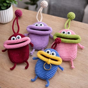 Op de afbeelding: Een verzameling van vijf handgemaakte gehaakte monsterzakjes in verschillende kleuren. Elk zakje heeft een brede, open mond met wiebelogen, kleine armen en benen. De zakjes zijn in rood, paars, roze, blauw en groen, met een lus en een bal aan de bovenkant.