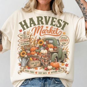 Könnte beinhalten: Ein cremefarbenes T-Shirt mit einer Grafik eines grünen Oldtimer-Trucks, beladen mit Kürbissen, Sonnenblumen und einer karierten Decke. Der Text lautet HARVEST Market, FALL, FLOWERS, PUMPKINS, LOCALLY GROWN, FARM FRESH, GOOD VIBES, COZY DAYS, AUTUMN WAYS.