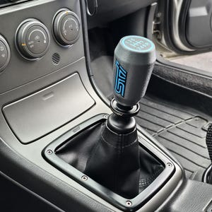 May include: A black and blue STI shift knob installed in a car. The shift knob has a blue gear pattern and the STI logo in blue. The shift boot is black leather, and the surrounding console is gray.