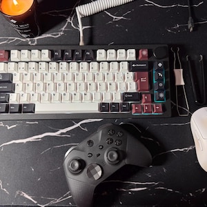 Può includere: Vista dall'alto di un'impostazione di gioco su una superficie nera con motivo marmorizzato. L'impostazione include una tastiera meccanica con tasti bianchi e bordeaux, un gamepad nero e un mouse wireless bianco. Una candela accesa è visibile.