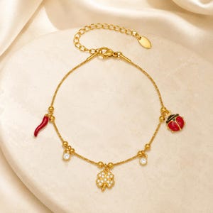 May include: A gold-tone chain bracelet featuring a red chili pepper charm, a red and black ladybug charm, two small clear round crystal charms, and a central four-leaf clover charm encrusted with small clear crystals, with an adjustable clasp.
