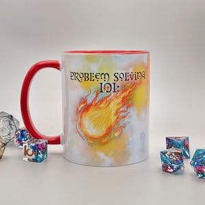 May include: A white ceramic mug with a red handle and interior rim. The front features a graphic of a flaming fireball and the black text "PROBLEM SOLVING 101:". Several multi-colored, translucent polyhedral dice are scattered around the base.