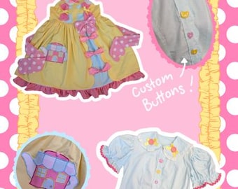 May include: A yellow dress with pink polka dot accents and a teapot graphic, a light blue short-sleeve shirt with a ruffled collar, and a circular inset showing small bear-shaped buttons. The text reads Custom Buttons! and TeaHouse.