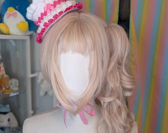 May include: A mannequin head with light blonde hair styled in pigtails and bangs, wearing a miniature cake-shaped hat. The hat is white with pink accents and a small candle. A pink ribbon is tied around the neck.