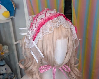 May include: A pink and white lace bonnet with a gingham ribbon bow. The bonnet has a pink band and white lace trim. White ribbons are attached. The item is displayed on a mannequin head with blonde hair.