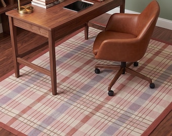 Puede incluir: Una alfombra rectangular con un patrón de cuadros en tonos rosa, morado claro y crema con un borde sólido de color terracota. La alfombra está colocada debajo de un escritorio de madera y una silla de oficina de cuero marrón con ruedas.