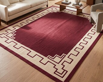 Puede incluir: Una alfombra rectangular grande con un campo central de color burdeos intenso rodeado por un borde ancho de color crema. El borde está decorado con un patrón geométrico continuo de estilo griego en el mismo color burdeos.