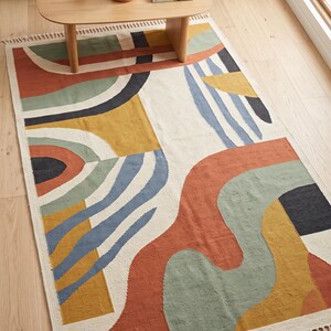 Puede incluir: Una alfombra rectangular con base color blanco roto y un patr&oacute;n abstracto multicolor de formas en terracota, verde salvia, amarillo mostaza y azul marino, que incluye l&iacute;neas curvas y bloques geom&eacute;tricos, rematada con flecos color crema.