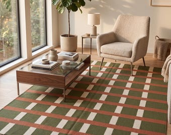 Puede incluir: Una alfombra rectangular con un patrón de cuadrícula de rayas en verde oliva, naranja terracota y blanco crema. La alfombra está colocada sobre un suelo de madera clara, extendiéndose desde el primer plano hacia una mesa de centro y un sillón.