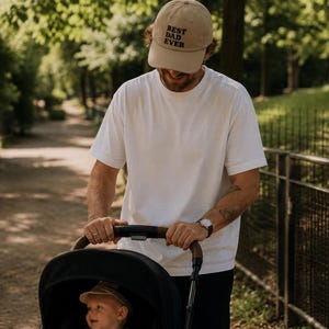 Peut inclure: Un chapeau de baseball beige avec le texte "BEST DAD EVER" en noir est porté par une personne portant un t-shirt blanc. La personne pousse une poussette noire avec un bébé à l'intérieur. La scène se déroule en extérieur dans un parc.