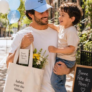 Peut inclure: Un homme porte une casquette bleu clair avec l'inscription "World's Greatest Dad". Il tient un enfant et un sac fourre-tout avec des fleurs et le texte "habibi trust the timing". Un panneau indique "Bonne Fête des Pères!"