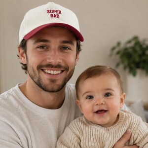 May include: A man wearing a white and maroon baseball cap with the text "SUPER DAD" smiles while holding a baby. The baby is wearing a cream-coloured sweater and smiling. The background includes a plant in a white pot.