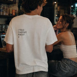 Peut inclure: Un t-shirt blanc avec le texte "I TOLD CHATGPT ABOUT U" imprimé au dos. Le t-shirt est porté par une personne dans un bar, en interaction avec une autre personne. La scène se déroule dans un bar faiblement éclairé.