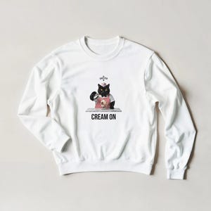Op de afbeelding: Een wit sweatshirt met lange mouwen en een afbeelding van een zwarte kat met een wit mutsje en een rood-wit gestreept schort. De kat houdt een klein kannetje boven een kom. De tekst CHESTER en CREAM ON is in het zwart gedrukt.