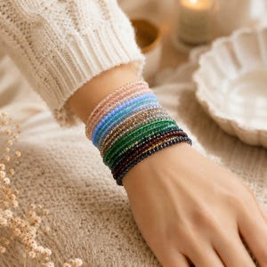 Può includere: Una serie di braccialetti sottili composti da piccole perline sfaccettate e traslucide in un gradiente di colori che include rosa chiaro, blu, verde, marrone e blu scuro, indossati insieme al polso per creare un aspetto a strati.