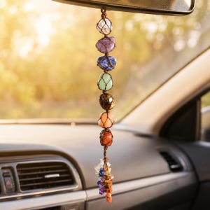 Puede incluir: Un adorno colgante para coche con siete piedras pulidas multicolores dispuestas verticalmente en una red de cordón marrón, incluyendo piedras transparentes, moradas, azules, verdes, marrones, naranjas y rojas, terminando en un racimo de cristales.