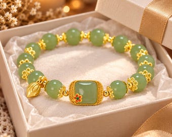 May include: A beaded bracelet featuring round light green jade-like stones and gold-toned metal spacers. A central rectangular green stone is accented with a small red and green flower detail, and a gold-toned leaf-shaped charm hangs from the side.