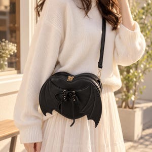 Può includere: Una borsa a tracolla nera a forma di cuore con ali di pipistrello decorative sui lati e un dettaglio centrale con lacci neri. La borsa presenta una finitura liscia, cerniere color oro e una sottile tracolla nera fissata sulla parte superiore.