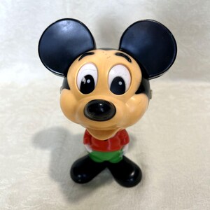 May include: A plastic figurine of a cartoon mouse character with large round black ears, a peach-colored face, a black nose, and white eyes. The character wears a red shirt, green pants, and black shoes, standing in a forward-facing position.