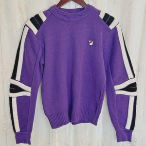 May include: A purple long-sleeve knit sweater featuring a crew neckline and ribbed cuffs. The sleeves are accented with vertical white and black stripes running from the shoulder to the cuff. A small Fila logo patch is on the upper left chest.