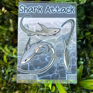 May include: A rectangular sticker sheet titled Shark Attack featuring three grey shark illustrations swimming in water. The design includes the text ADUBROV and 1100 at the bottom. The sharks are depicted with long, curved tails and detailed fins against a blue water background.