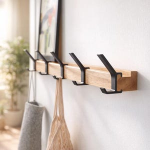 Peut inclure: Un porte-manteau mural en bois avec cinq crochets en métal noir. Le support est en bois clair et est fixé à un mur blanc. Deux sacs sont suspendus aux crochets. Une image encadrée est au-dessus.
