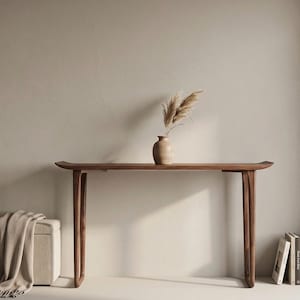Könnte beinhalten: Eine Konsole aus dunklem Holz mit geschwungener Oberseite und Beinen. Eine beige Vase mit getrocknetem Gras steht auf dem Tisch. Eine beige Aufbewahrungs-Ottomane und ein Stapel Bücher befinden sich auf beiden Seiten. Der Tisch ist etwa 183 cm lang.