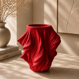 May include: A striking red ceramic vase featuring a draped fabric design. The vase has a wide opening and a textured, flowing appearance, adding a modern touch to home decor. The vase is displayed on a light-coloured surface.