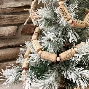 May include: A close-up of a Christmas tree with a garland made of wine corks. The corks are strung together with wooden beads. The tree has green needles and is dusted with white snow. Some corks have text such as "COPPOLA" and "COLUMBIA VALLEY".