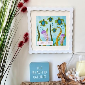 May include: A square white scalloped frame containing an embroidered artwork of sea turtles and jellyfish in blue water with green seaweed. A small white label at the bottom features the black text "go with the flow" in a typewriter font.
