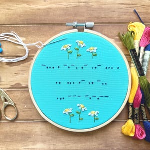 May include: A circular wooden embroidery hoop containing bright turquoise fabric with hand-stitched white daisy flowers and dark blue Morse code patterns. Beside the hoop are silver stork-shaped scissors, white thread, and several colorful embroidery floss skeins.