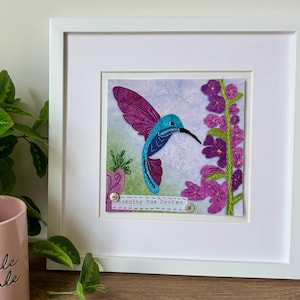 Peut inclure: Une œuvre d'art textile encadrée représentant un colibri bleu et violet vibrant près d'une tige de fleurs violettes. L'œuvre comprend une petite étiquette en tissu avec le texte brodé "morning has broken" fixé par deux petits boutons.