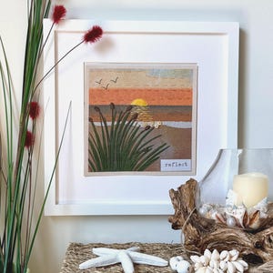 May include: A square framed textile art piece featuring an embroidered sunset over a beach with tall green grasses in the foreground. The scene includes a yellow sun, orange sky, and two birds. The word reflect is printed in a small white box.