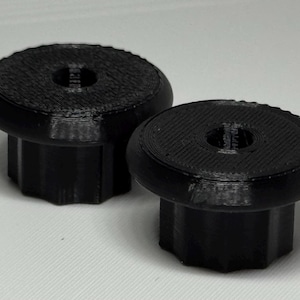 May include: Two black, circular knobs with a textured top surface and a central hole. Each knob features a fluted, gear-like base with vertical ridges, designed for gripping and turning. The objects have a matte finish and a uniform black colour.