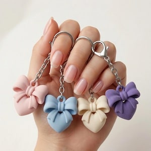 May include: Four heart-shaped keychains featuring matte bows in pastel pink, light blue, cream, and purple. Each charm is attached to a silver-toned metal chain and a lobster clasp, showcasing a smooth, solid-colored finish on each heart and bow.