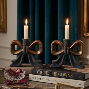 May include: Two matte charcoal grey candle holders shaped like bows with metallic bronze accents, each holding a single cream-coloured taper candle. The bow-shaped candle holders are positioned on top of stacked hardcover books.
