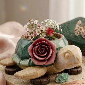 May include: A turtle-shaped planter with a faceted green shell, featuring a large dark pink rose attached to the front. The planter holds a small arrangement of white and pink flowers. The turtle has a light beige head and dark brown feet.