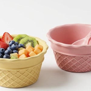 May include: A yellow bowl filled with sliced strawberries, blueberries, kiwi, and cantaloupe, alongside a pink bowl with a matching pink spoon. Both bowls feature a textured waffle-cone pattern on the exterior and a scalloped rim design.