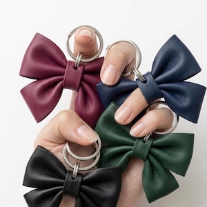 Puede incluir: Una colección de cuatro llaveros con forma de lazo hechos de cuero liso en colores burdeos, azul marino, verde bosque y negro. Cada lazo cuenta con una banda central de cuero y está unido a una anilla metálica plateada para las llaves.
