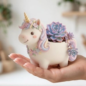 Könnte beinhalten: Ein cremefarbener Einhorn-Pflanztopf mit goldenem Horn, pastellfarbener Mähne und floralen Details. Er enthält eine blau-grüne Sukkulente. Ein bezauberndes Dekorationsstück.