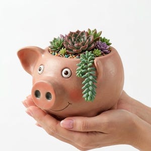 May include: A ceramic pig-shaped planter in a light pink hue, filled with a variety of succulents and small decorative stones. The planter has a smiling face and is held in two hands.