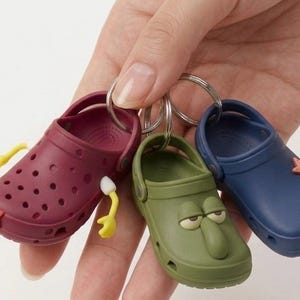 May include: Three miniature Crocs keychains in burgundy, green, and navy blue. The burgundy shoe has yellow and white accents, the green one has a face, and the navy blue one has a starfish charm. Each keychain is attached to a silver ring.