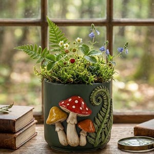 May include: A dark green cylindrical ceramic planter featuring three-dimensional relief decorations of a large red-capped mushroom with white spots, two smaller yellow and orange mushrooms, and a green fern frond, filled with green moss and plants.