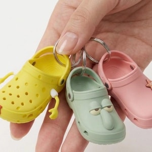 May include: A set of three miniature shoe-shaped keychains in yellow, mint green, and pink. The yellow keychain has arms and a red detail, the green one has a face, and the pink one has a starfish. All three are attached to a silver keyring.