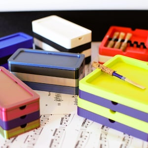 Peut inclure: Plusieurs boîtes rectangulaires colorées, bleues, rouges, vertes et violettes. Certaines sont empilées, et l'une contient des anches. Une anche est posée sur une boîte jaune et violette. Des notes de musique sont en arrière-plan.